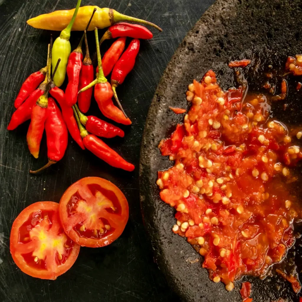 Sambal dalam cobek batu, cabai merah, dan irisan tomat.