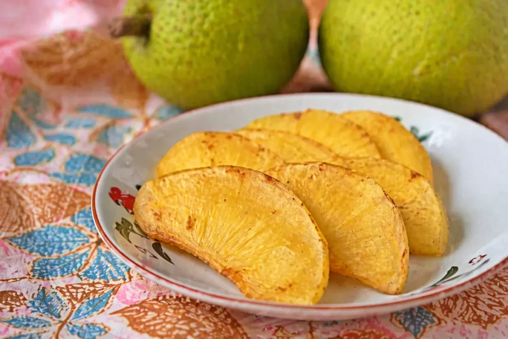 Sukun goreng disajikan di atas piring dengan latar belakang buah sukun yang masih utuh.