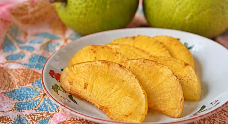 Sukun goreng disajikan di atas piring dengan latar belakang buah sukun yang masih utuh.
